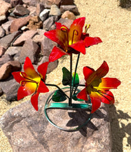 Load image into Gallery viewer, A metal garden sculpture of red lilies with a yellow center on a green wire stand, placed outdoors on a rock with rocks and sand in the background.
