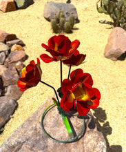 Load image into Gallery viewer, Red metal garden flowers with yellow centers mounted on a green circular wire stand, placed outdoors on a rock in a sandy desert garden with cacti in the background.
