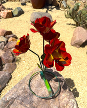 Load image into Gallery viewer, Three red metal garden flowers with yellow and orange accents, mounted on a green base, displayed outdoors on a stone in a sandy desert garden setting.
